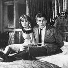 Es gibt keinen Text. Zwei Personen sitzen überrascht auf einem Bett, in einem Raum mit Fenstern und Vorhängen.