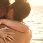 Eine Frau und ein Mann umarmen sich innig am Meer, beleuchtet von warmem Sonnenlicht.