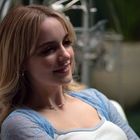 Eine Frau lächelt, sitzt in einem hellen Raum. Sie trägt ein blaues Spitzentop. Im Hintergrund sind unscharfe Pflanzen zu sehen.