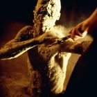 Eine von Staub und Erde bedeckte, menschliche Figur erhebt sich, während eine Hand eine Schere in der Nähe hält.
