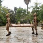 Zwei Personen in Khaki-Uniformen tragen eine Trage durch einen regnerischen, von Bäumen und einem Gebäude gesäumten Innenhof.