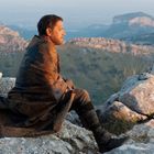 Ein Mann in mittlerem Alter sitzt auf felsigem Terrain, in Gedanken versunken, mit Bergen im Hintergrund.