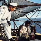 Ein weiß gekleideter Seemann steht auf einem Schiff, nachdenklich auf einen sitzenden Jungen blickend, umgeben von Netzen und Meer.