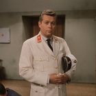 Ein Mann in weißer Uniform mit roten Abzeichen, hält eine weiße Mütze. Im Hintergrund, ein Erste-Hilfe-Kasten an der Wand.