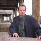 Ein Mann in dunklem Mantel steht nachdenklich an einem Holzkai, im Hintergrund Holzhütten und Wasser.