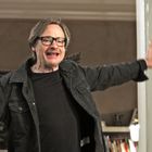 Ein Mann in Brille und Jacke steht lächelnd mit offenen Armen in einem Raum mit geöffneter Tür.