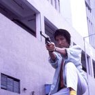 Ein Mensch kniet mit ausgestrecktem Arm und hält eine Pistole. Er schaut konzentriert, im Hintergrund ein Gebäude.