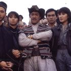 Fünf Menschen stehen eng beieinander, alle schauen ernst. Im Hintergrund sind weitere Personen zu sehen.