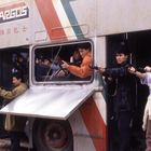 „ARGOS“-Logo, chinesische Schrift. Männer mit Pistolen lehnen aus altem Doppeldeckerbus in urbaner Umgebung.