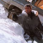 Ein Mann in einem dicken Parka zielt mit einer Pistole, teilweise verschneit, steht er vor einem geneigten Fahrzeug.