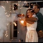 Ein Mann und eine Frau stehen eng beieinander, während sie eine Waffe abfeuern. Funken fliegen um sie herum.