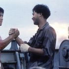 Zwei Männer schütteln sich die Hände auf einem Schiff. Einer trägt einen blutverschmierten Tanktop, der andere ein dunkles Hemd.