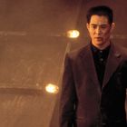 Kein Text vorhanden. Ein Mann in einem braunen Anzug steht vor einem verschwommenen Hintergrund mit leuchtenden Lichtern.