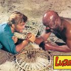 Text "Luana" in gelber Box. Zwei Männer im Sand, Händedrücken, lachen, einer trägt blau, der andere ist oberkörperfrei.