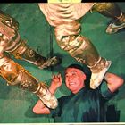 Ein Mann liegt am Boden, lächelt und hebt die Hände. Über ihm stehen zwei Personen in goldenen Kostümen.