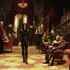 Keine sichtbaren Texte. Eine Frau in schwarzem Anzug schreitet durch einen opulenten Raum voller vornehm gekleideter Personen.