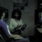 Zwei Personen unterhalten sich in einem Raum, während ein Mann auf einem Fernseher im Hintergrund spricht.