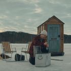 Text: "OFFICE". Zwei Personen am See auf Eis; einer öffnet ein Getränk, der andere hält einen Eisbohrer neben einer Hütte.