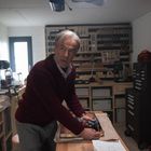Ein älterer Mann in einer Werkstatt hält eine Bohrmaschine. Umgeben von Regalen voller Werkzeuge und Schubladen.