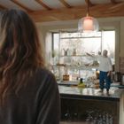 Eine Person mit langem Haar steht in einer Küche, betrachtet eine Miniaturfigur auf der Arbeitsplatte.