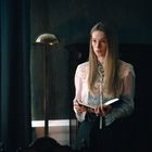 Eine junge Frau mit langen blonden Haaren hält ein Buch. Sie trägt eine weiße, verzierte Bluse und steht in einem dunklen Raum.