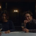 Zwei Personen sitzen an einem Tisch mit Weingläsern. Die Atmosphäre ist gedämpft, warmes Licht scheint von Wandleuchten.