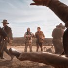 Western-Szene mit Cowboys im Kampf, zwei Personen in traditionellen Kleidern beobachten im Hintergrund. Landschaftlich trocken.
