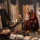 Menschen in mittelalterlicher Kleidung in einem Raum mit hängenden Stoffbahnen, Werkzeugen und einer brennenden Kerze.