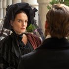 Ein Mann und eine Frau in altmodischer Kleidung sitzen sich gegenüber in einer Säulenhalle. Die Frau trägt einen schwarzen Hut mit Schleier.