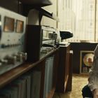 Ein Mann mit Kopfhörern sitzt konzentriert vor Audiogeräten und Schallplatten, während im Hintergrund große Fenster sind.