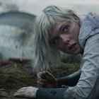 Eine Frau mit blonden Haaren liegt erschöpft auf dem Boden, umgeben von einer grasigen Landschaft.