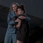 Zwei Personen umarmen sich, eine davon hält einen kleinen Hund. Es scheint Nacht zu sein, sie wirken besorgt.