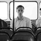 Drei Personen sitzen in einem Bus. Links sitzt ein Mann mit gesenktem Kopf, in der Mitte ein Mann frontal, rechts eine Frau.