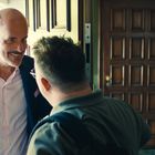 Zwei Männer an einer Haustür, lächelnd. Einer mittleren Alters im Anzug, der andere mit Brille und Rucksack. Im Hintergrund eine Landkarte.