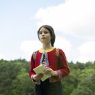 Ein Kind im bunten Cardigan hält ein Notizbuch und steht vor einem Wald, der Himmel ist blau mit ein paar Wolken.