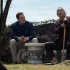 Ich kann den Text in diesem Bild nicht erkennen. Zwei Männer sitzen im Freien auf Steinhockern, einer hält einen Stock.