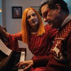 Notenblätter auf dem Klavier; eine Frau mit rotem Haar beobachtet einen Mann beim Spielen. Beide in roter Kleidung.