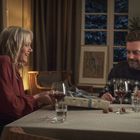 Ein Paar sitzt an einem Tisch mit Weingläsern. Die Frau hält ein Geschenk. Im Hintergrund ein Fenster und Blumenschmuck.