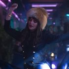 Eine Frau tanzt ausgelassen in einem Club, trägt Bärenfellmütze und schwarzen Mantel. Die Umgebung ist dunkel mit bunten Lichtern.