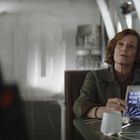 Eine Frau sitzt an einem Tisch in einem futuristischen Raum und betrachtet ein holografisches Display.