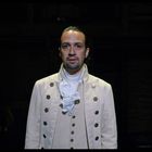 Ein Mann in historischer Kleidung mit weißem Rüschenhemd steht im Spotlight auf einer dunklen Bühne.
