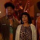 Text "Roll Up" auf einer Jacke. Drei Personen wirken überrascht: links ein Mann mit Afro, in der Mitte eine Frau mit Leoparden-Muster und rechts ein Cowboy.