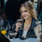 Eine Frau mit blonden Haaren hält ein Cocktailglas. Sie trägt eine schwarze Lederjacke. Im Hintergrund ist eine weitere Person.