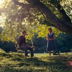 Zwei Personen unter einem großen Baum, eine Person sitzt auf einer Schaukel, die Sonne strahlt durch die Blätter.