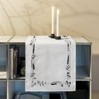 Ein Regal mit Büchern, Kerzenhaltern und einem weißen Tuch mit floralen Mustern. Schatten von Pflanzen an der Wand.