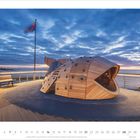 Kalender zeigt November 2027. Außerdekoration mit Holzstruktur in Fischform, Bank und Fahne bei Sonnenuntergang an der Küste.