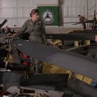 Grünes Banner mit Helikopter-Emblem im Hintergrund. Mehrere Personen in Fliegeranzügen reparieren Helikopter in einer Halle.