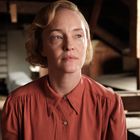 Ich kann keine Personen identifizieren. Eine Frau sitzt in einem Dachboden, trägt ein rotes Hemd, im Hintergrund Koffer.