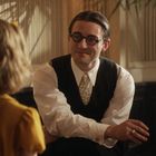 Ein Mann im Anzug und Brille sitzt lächelnd neben einem Klavier. Eine Frau in gelbem Kleid hört ihm aufmerksam zu.