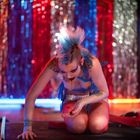 Eine Person mit blauem Irokesenschnitt und auffälligem Make-up kniet auf schwarzem Boden, umgeben von buntem, funkelndem Hintergrund.